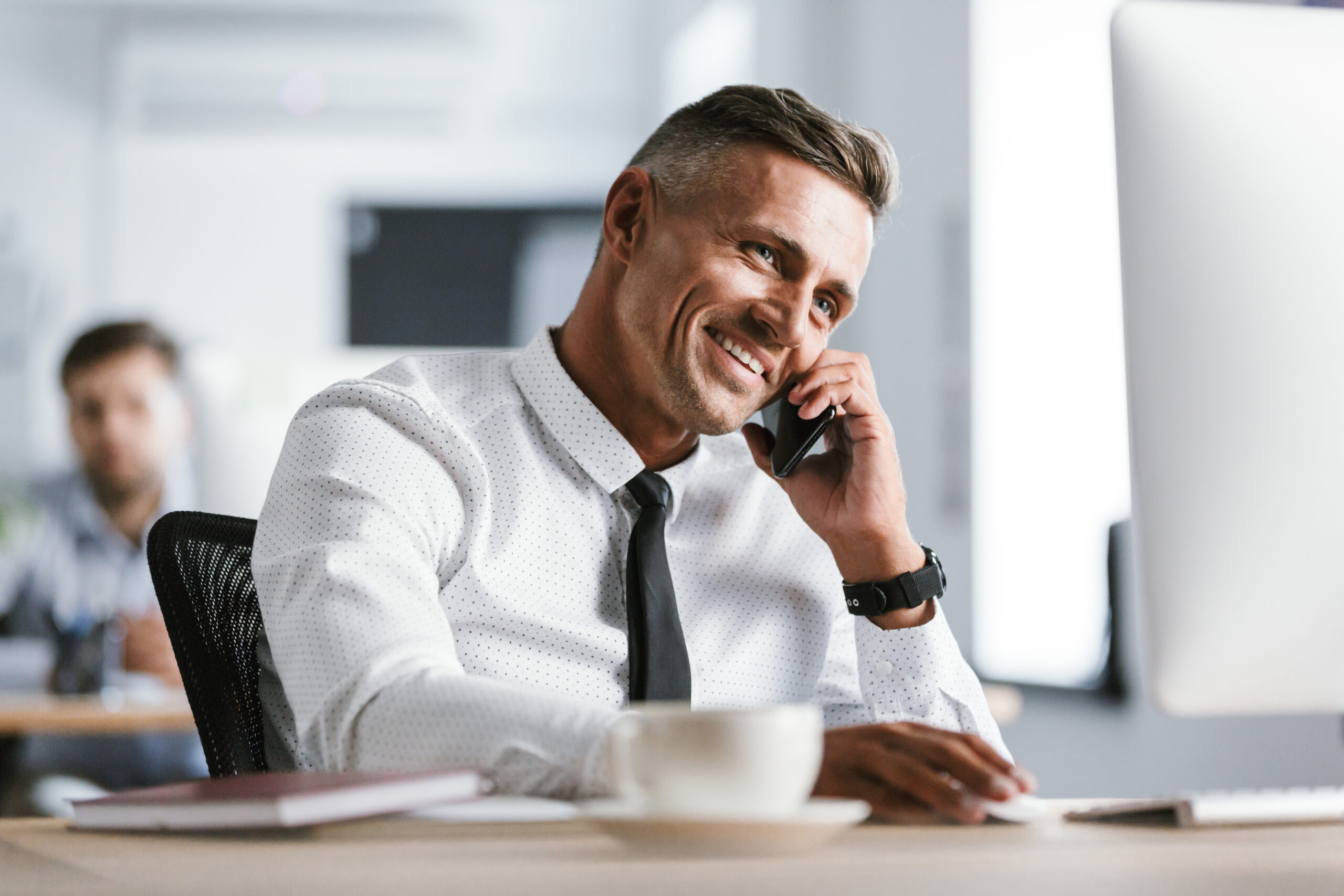 Image of European adult man 30s wearing white shirt and tie sitting at desk in office by computer and talking on smartphone